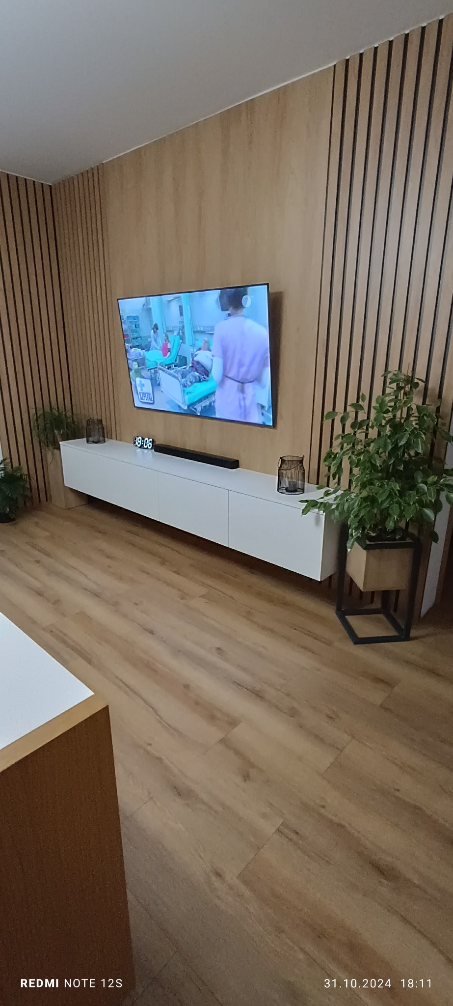 Biała szafka RTV pod telewizorem na tle ściany z drewnianych paneli. Na szafce dekoracje i zegar elektroniczny. Parkiet z paneli drewnopodobnych.