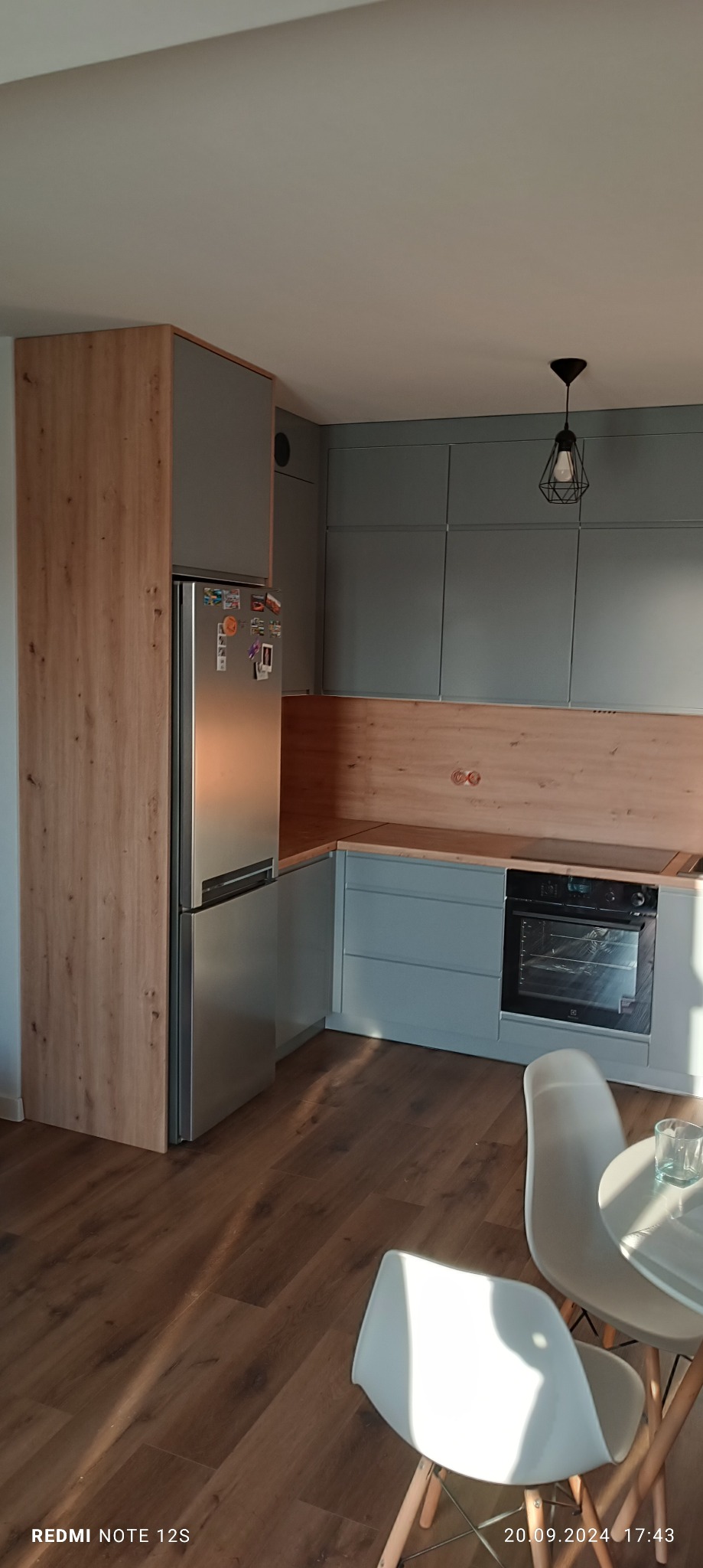 Szara kuchnia z drewnianymi akcentami: zabudowa lodówki, szafki wiszące i blat. Widoczny fragment stołu z krzesłami. Nowoczesny design, ciepłe oświetlenie.