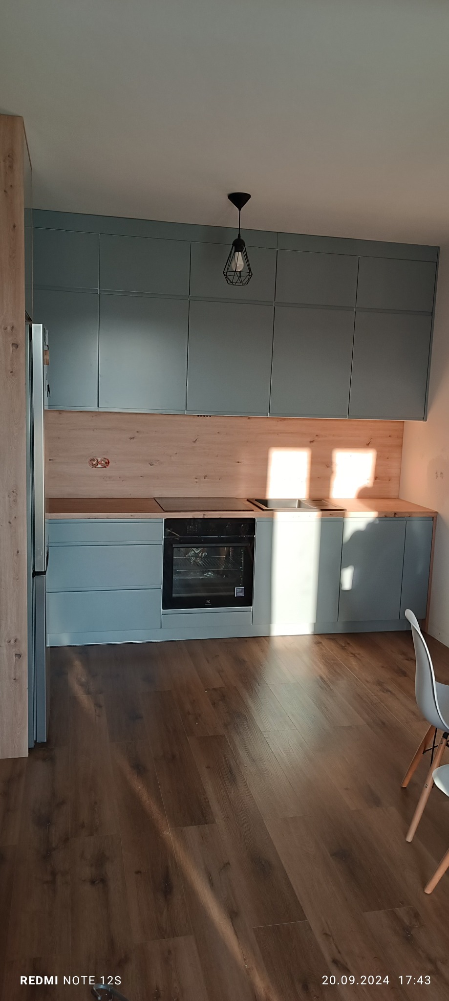 Kompletna kuchnia z szarymi szafkami i drewnianym blatem, oświetlona naturalnym światłem słonecznym, z widoczną lampą wiszącą w stylu industrialnym.