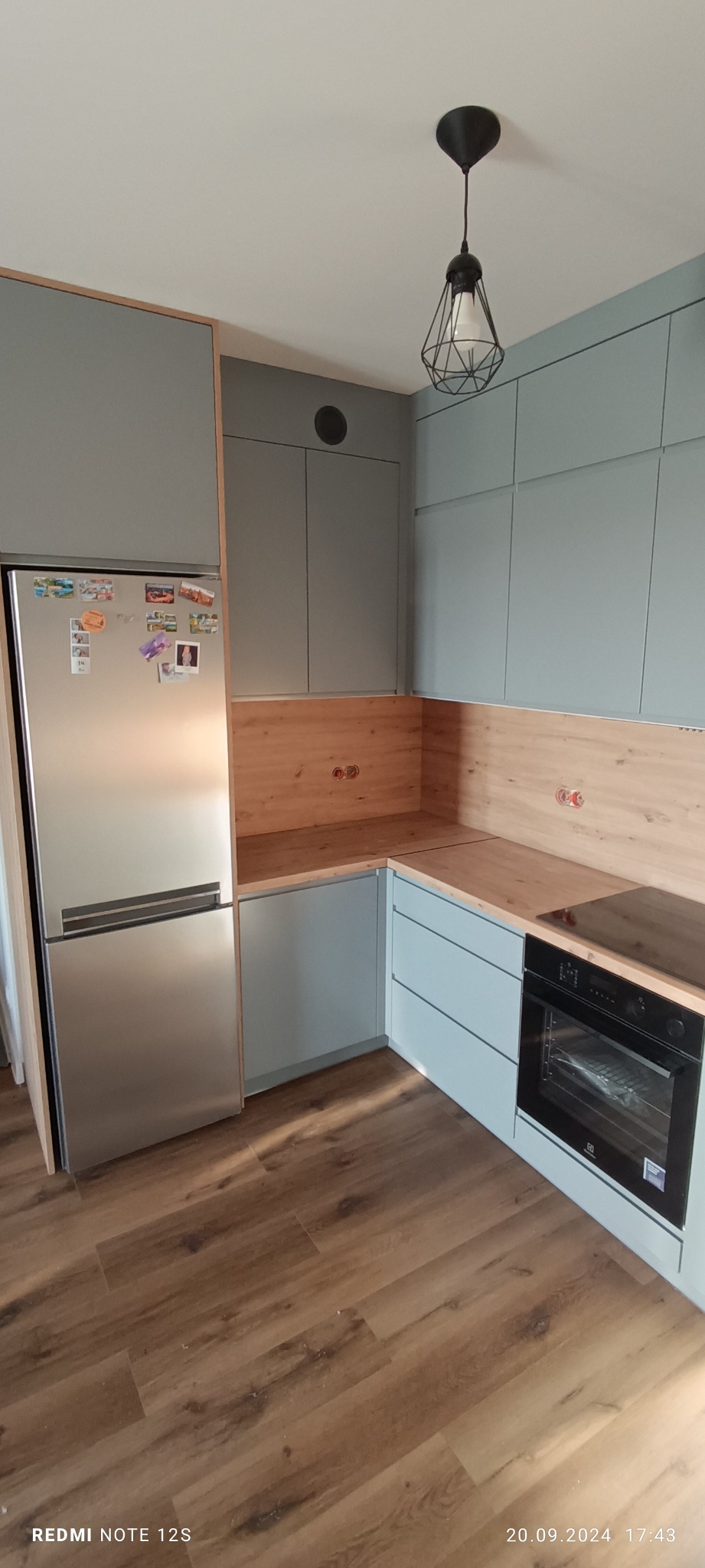 Narożna, szara kuchnia na wymiar z drewnianym blatem i srebrną lodówką. Nad blatem drewniana ściana. Wisząca lampa w stylu industrialnym. Całość w nowoczesnym stylu.