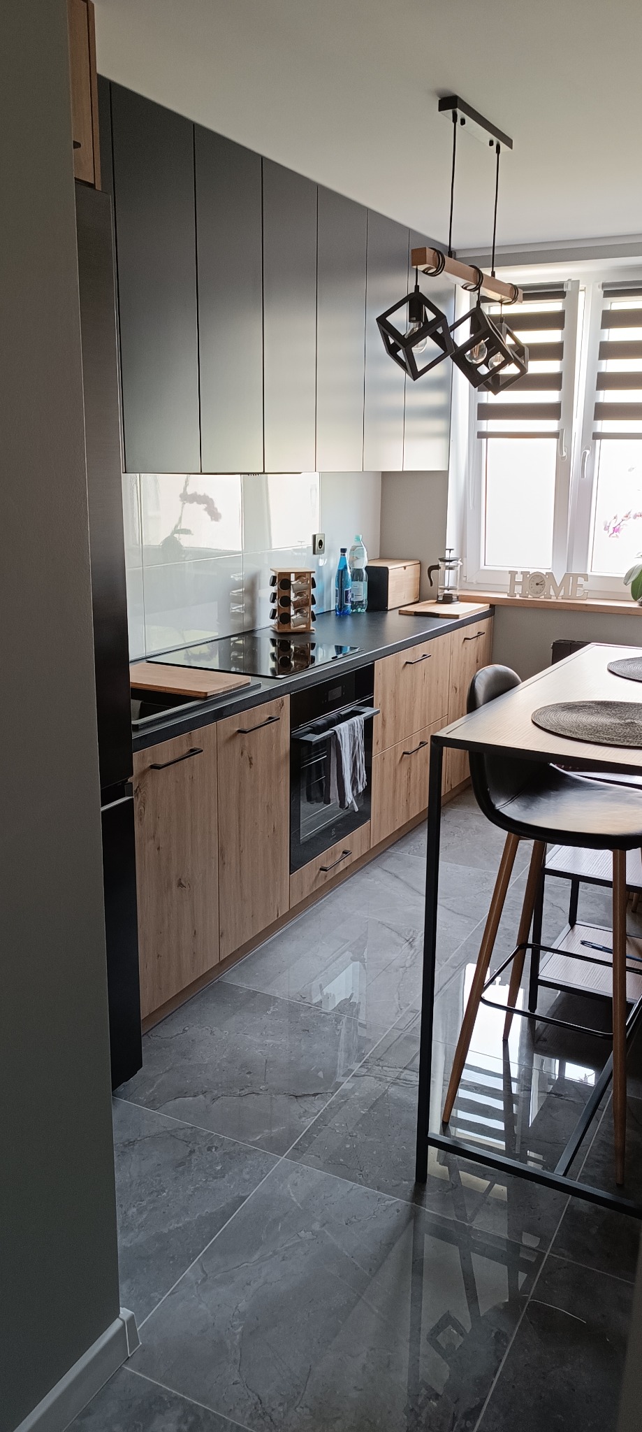 Nowoczesna kuchnia z szarymi szafkami górnymi i drewnopodobnymi dolnymi. Czarny blat, indukcja, piekarnik. Nad blatem białe płytki. Na podłodze szare płytki imitujące kamień.