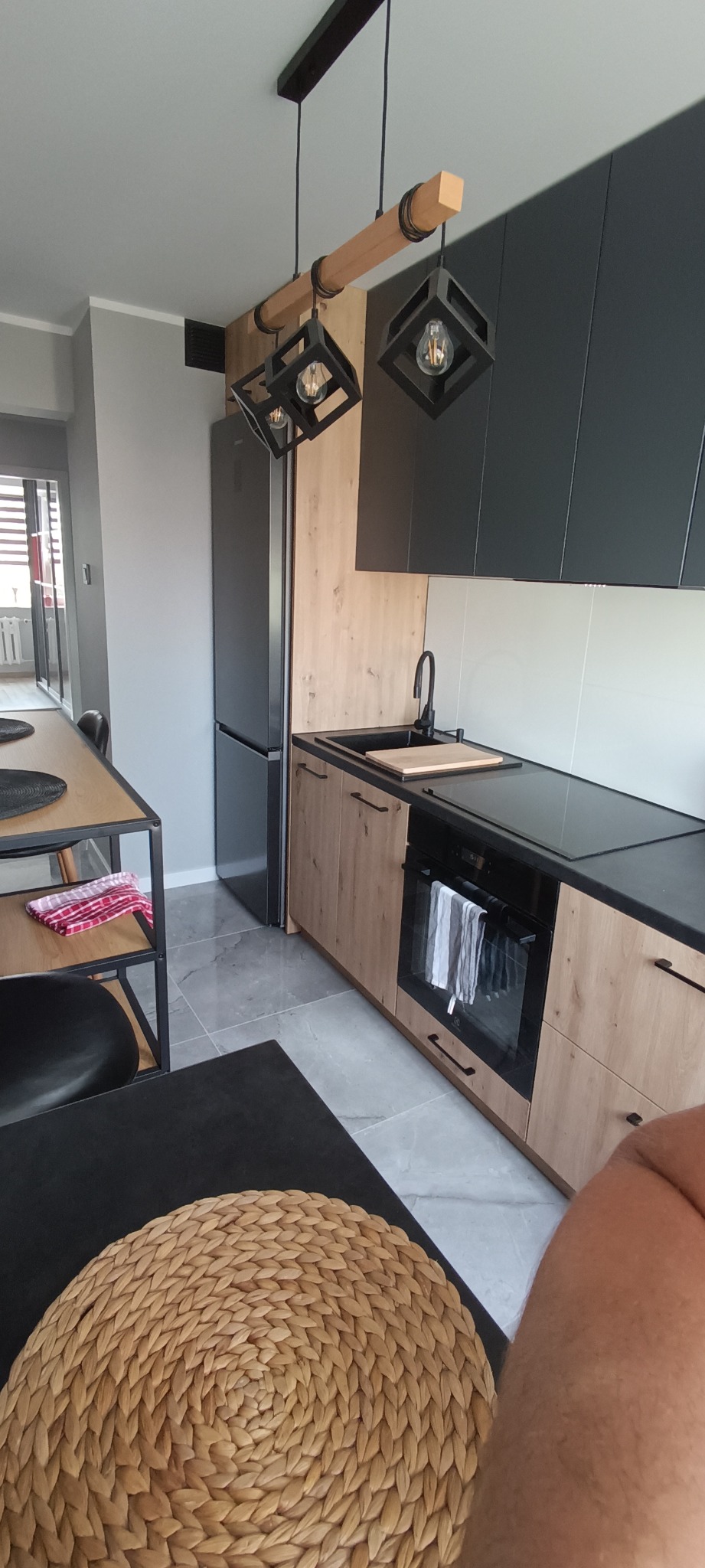 Nowoczesna kuchnia z drewnianymi szafkami dolnymi i czarnymi górnymi. Nietypowe lampy wiszące w stylu loft. Widoczny fragment stołu z plecioną podkładką.