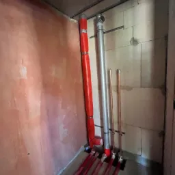 Nowa instalacja wodna i grzewcza w surowym pomieszczeniu: czerwone rury w izolacji, miedziane rury i srebrna rura wentylacyjna przy ścianach z betonu komórkowego i otynkowanej ścianie.