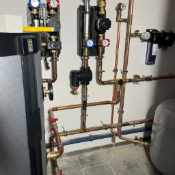 Kotłownia z powietrzną pompą ciepła Alpha Innotec, widoczne miedziane rury, liczniki, zawory i izolacje, zamontowane przy białej ścianie i szarej podłodze z płytek.