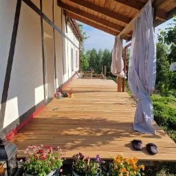 Nowy drewniany taras przy domu w stylu szachulcowym, z trzema donicami z kwiatami i zwiewnymi zasłonami.