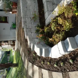 Kaskadowy murek z szarych bloczków betonowych tworzący rabaty w ogrodzie, z nowo posadzonymi roślinami i ścieżką z płyt. Widoczny fragment domu z donicami i konewką.