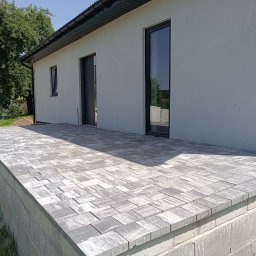 Sebastian Mitoraj Uslugi Brukarskie Perfekt-Bruk - Ukończony taras z szarej kostki brukowej o nieregularnym wzorze, przylegający do białego budynku z czarnymi oknami i drzwiami. W tle pracownik z taczką.