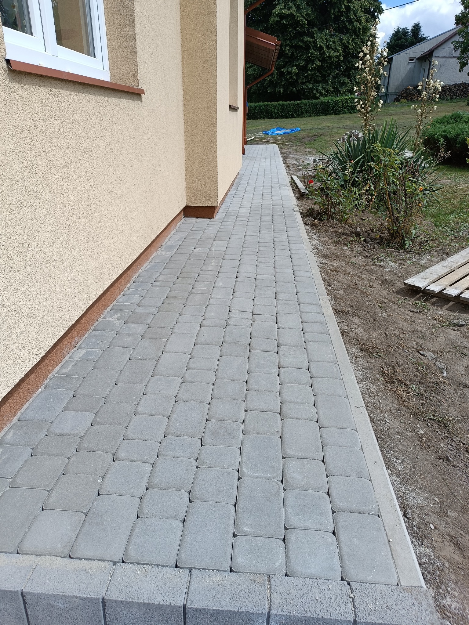 Szary chodnik z kostki brukowej ułożony wzdłuż ściany budynku z beżową elewacją, widoczne elementy ogrodu i nieba.