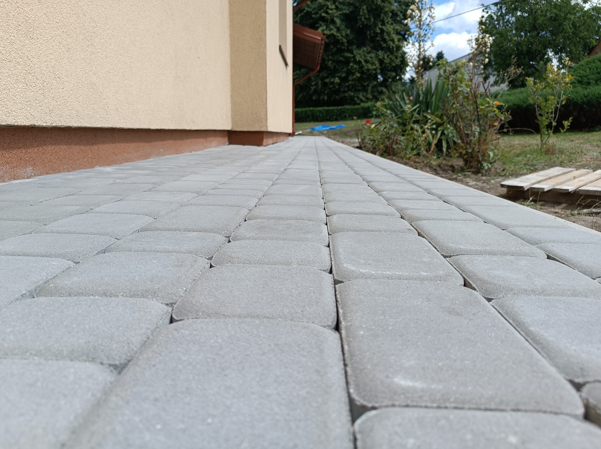 Świeżo ułożona, szara kostka brukowa przed budynkiem z beżową elewacją, widok z niskiej perspektywy, z fragmentami zieleni w tle.