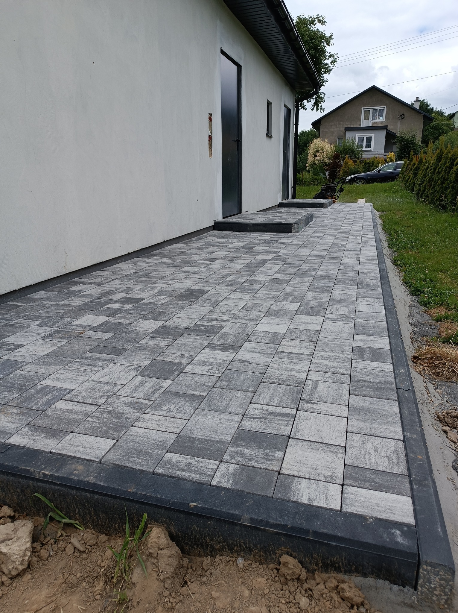 Ułożona szara kostka brukowa przed budynkiem z czarnymi drzwiami i stopniami, widok na ogród i dom w tle.