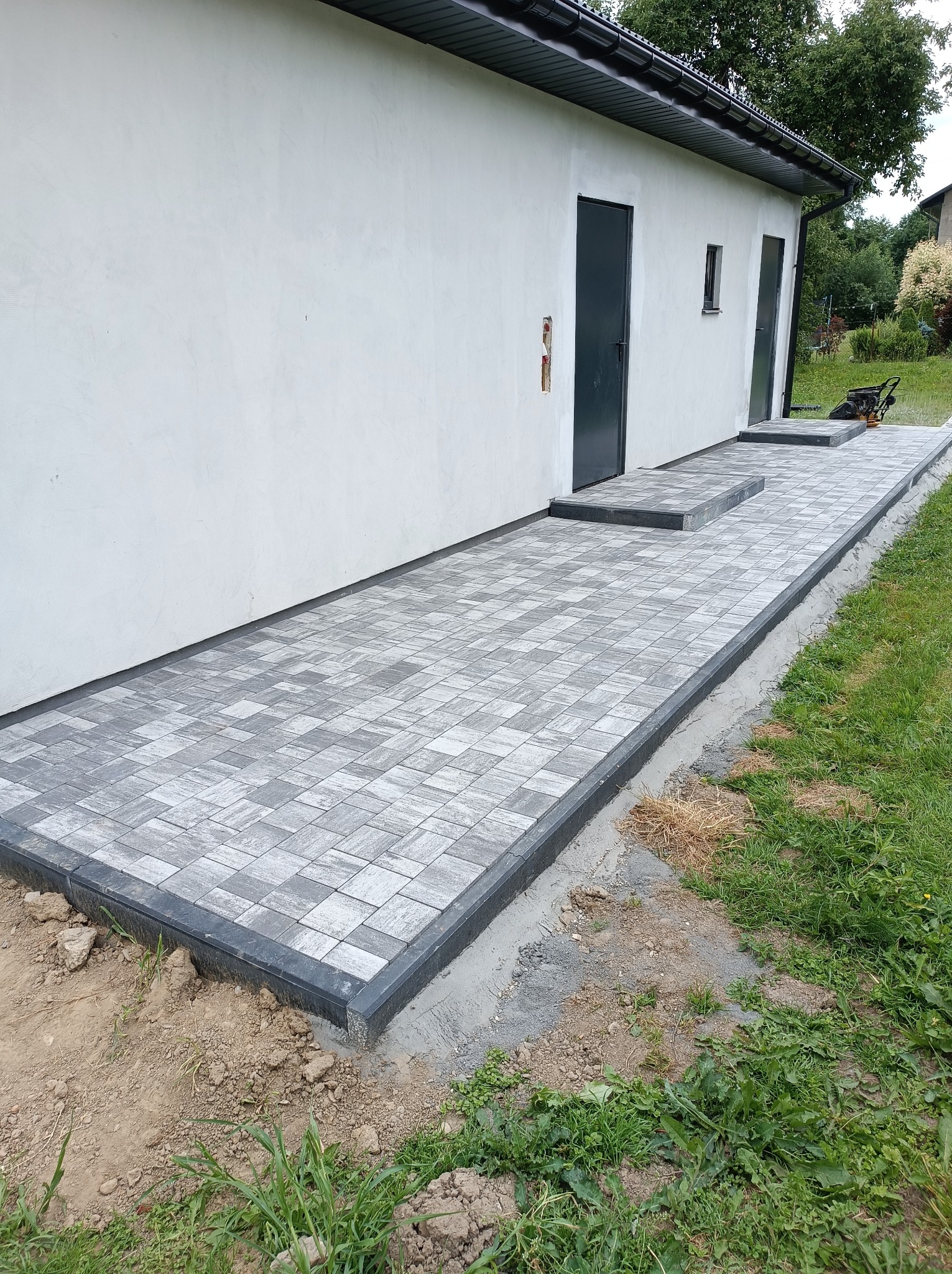 Świeżo ułożona szara kostka brukowa prowadząca do drzwi budynku, widoczna ubijarka w tle, trawa i ziemia wokół obrzeży.