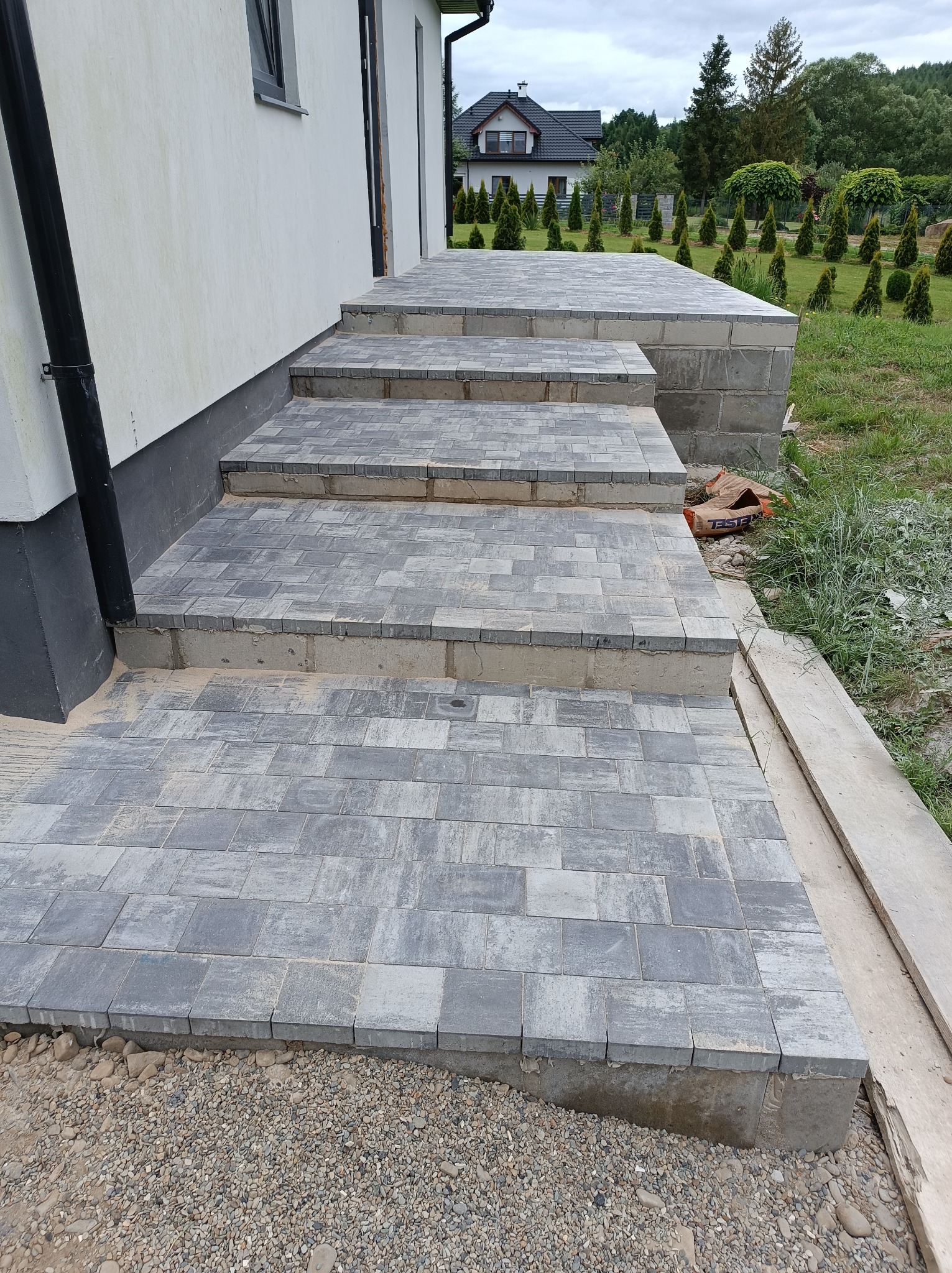 Schody wejściowe wyłożone szarą kostką brukową, widoczne worki z zaprawą, w tle dom z ogrodem.