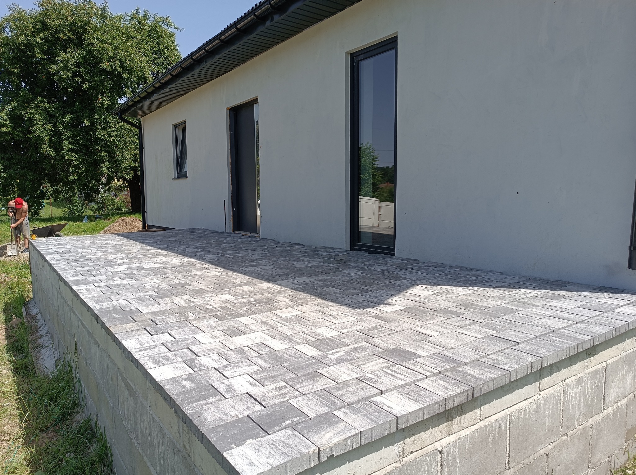 Ukończony taras z szarej kostki brukowej o nieregularnym wzorze, przylegający do białego budynku z czarnymi oknami i drzwiami. W tle pracownik z taczką.