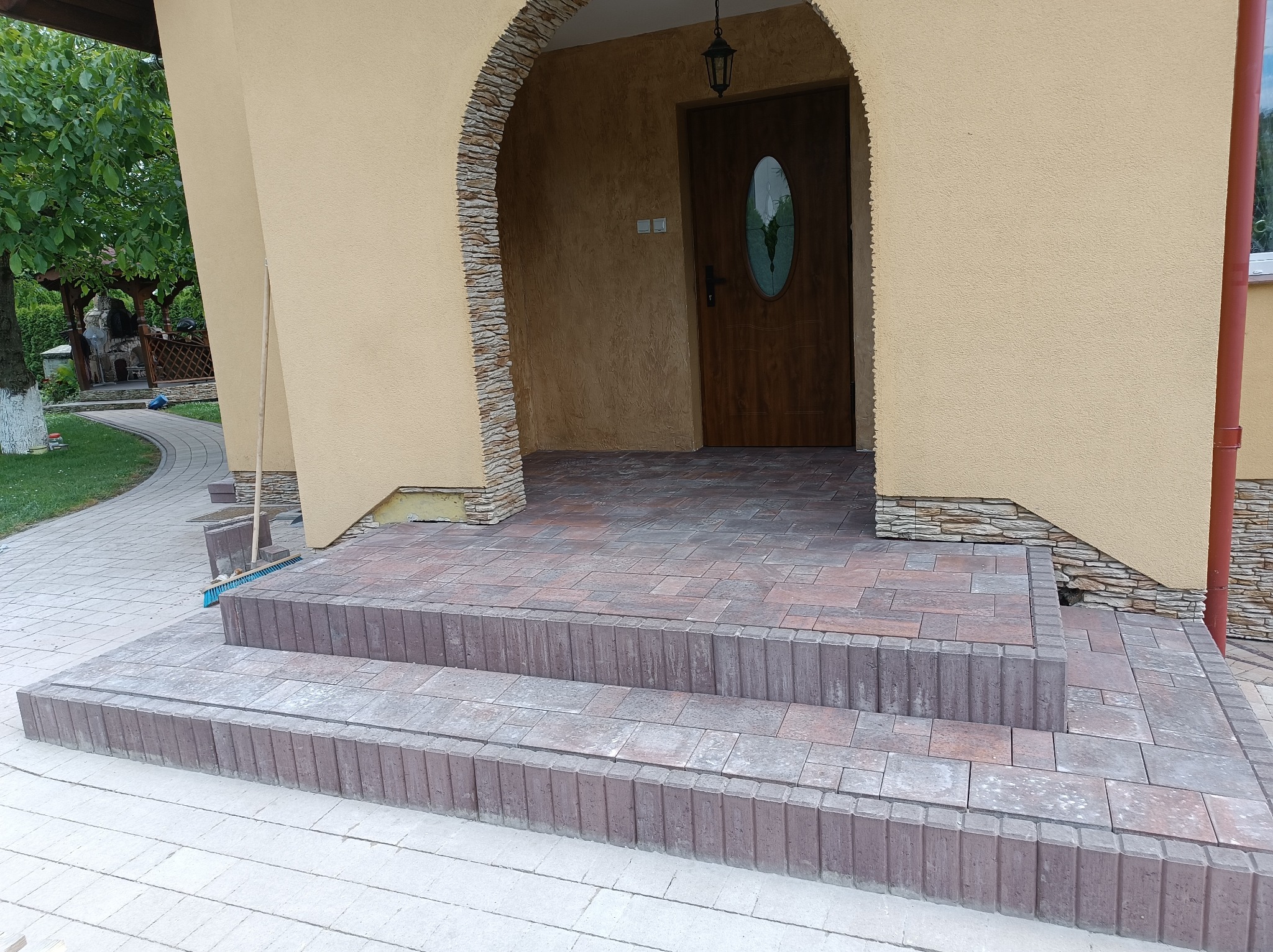 Dwu-stopniowe schody wejściowe wyłożone brązowo-szarą kostką brukową, prowadzące do domu z łukowym wejściem i drewnianymi drzwiami z owalnym przeszkleniem. Widoczny fragment chodnika z jasnoszarej...