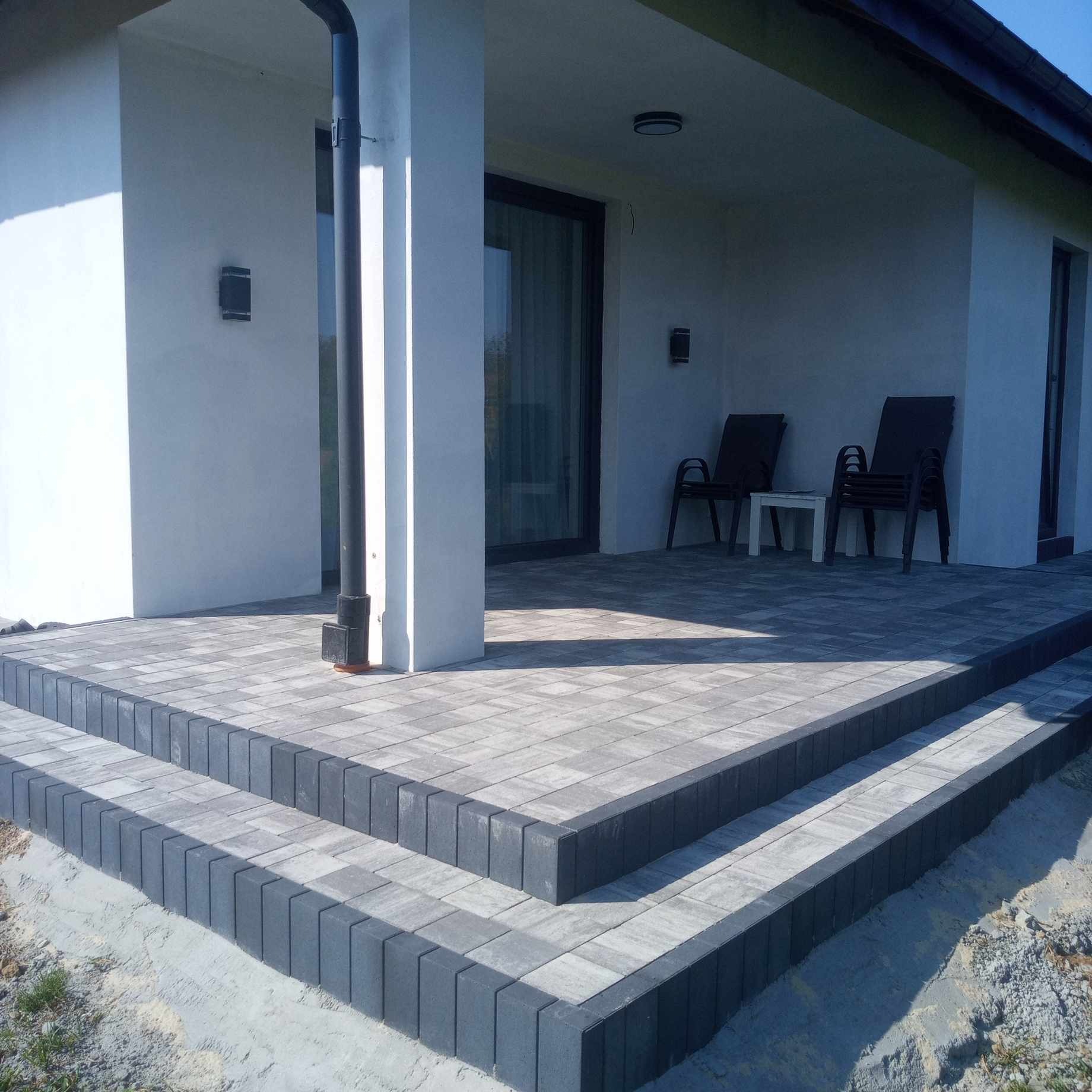 Wykończony taras z kostki brukowej w odcieniach szarości, z dwoma stopniami obrzeżonymi ciemniejszą kostką, przy białym budynku z czarnymi ramami okiennymi i drzwiami.