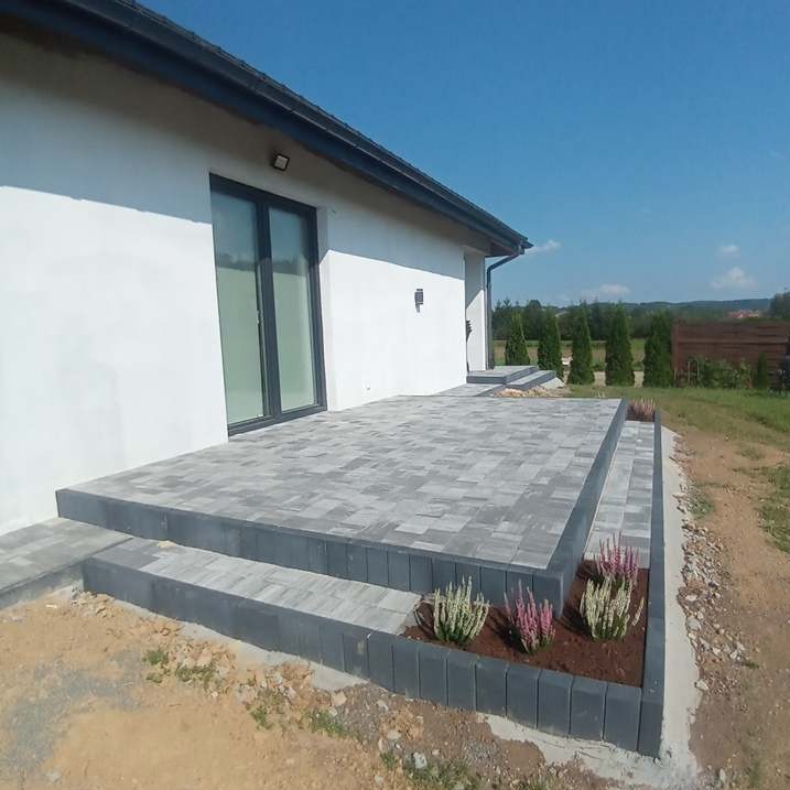 Nowoczesny taras z szarej kostki brukowej ułożony przed białym domem z ciemnymi oknami, rabata kwiatowa wzdłuż obrzeża, błękitne niebo w tle.