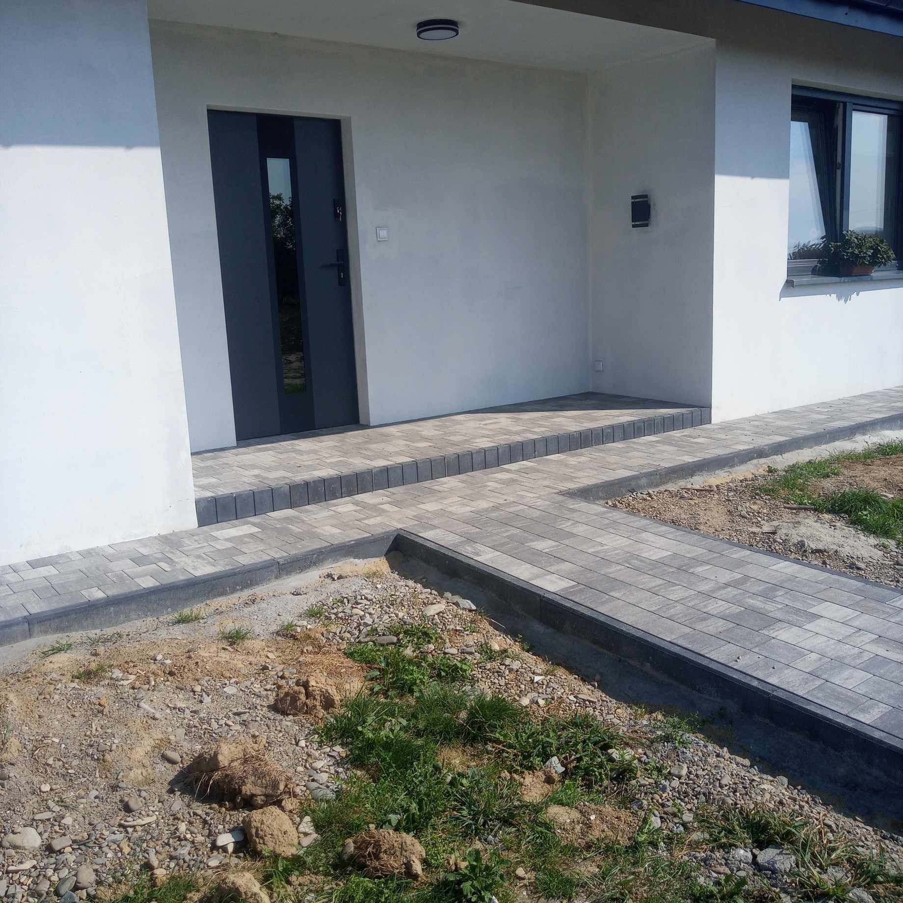 Nowoczesny chodnik z szarej kostki brukowej prowadzący do wejścia domu z ciemnymi drzwiami, widoczne niedokończone prace ziemne po bokach.