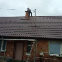 Pracownik na dachu z brązową dachówką, obok komina, montuje odgromnik, widoczne rusztowanie przy ceglanym budynku.