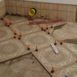 Układanie wzorzystych płytek podłogowych z użyciem pomarańczowych klipsów poziomujących, widoczna poziomica i rura kanalizacyjna w tle.