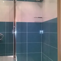 Wykończona łazienka z kabiną prysznicową z zaokrąglonym szkłem i turkusowymi płytkami do połowy ściany, powyżej białe płytki z turkusowym paskiem na górze, widoczne rury i włącznik na ścianie.