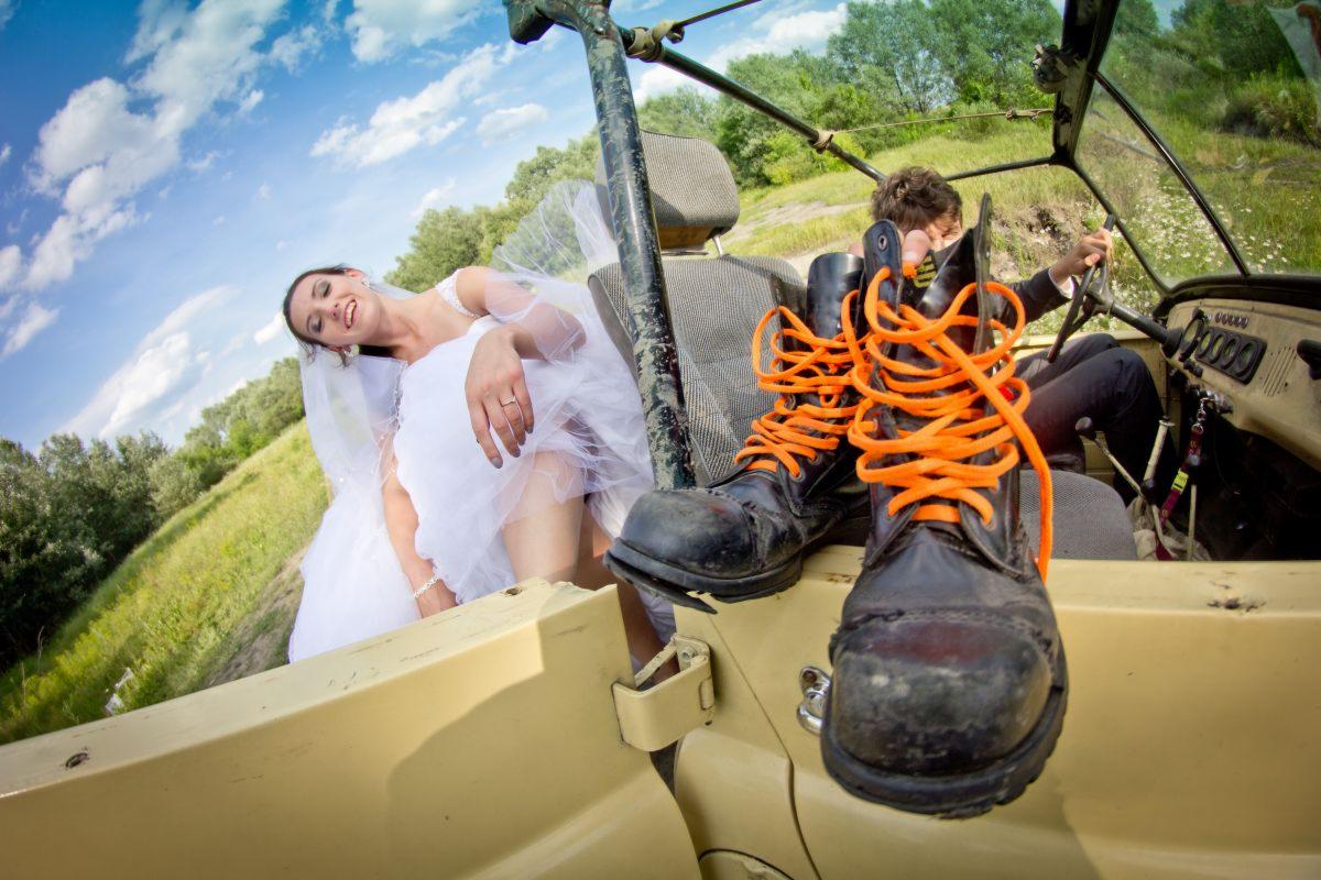 Panna młoda w białej sukni leży w jeepie, obok buty trekkingowe z pomarańczowymi sznurówkami, w tle kierowca i zielony krajobraz.
