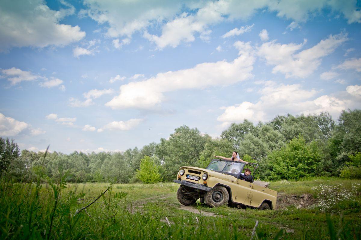 Para młoda w beżowym samochodzie terenowym bez dachu, na tle zieleni i błękitnego nieba z chmurami, panna młoda stoi z wyciągniętymi ramionami.