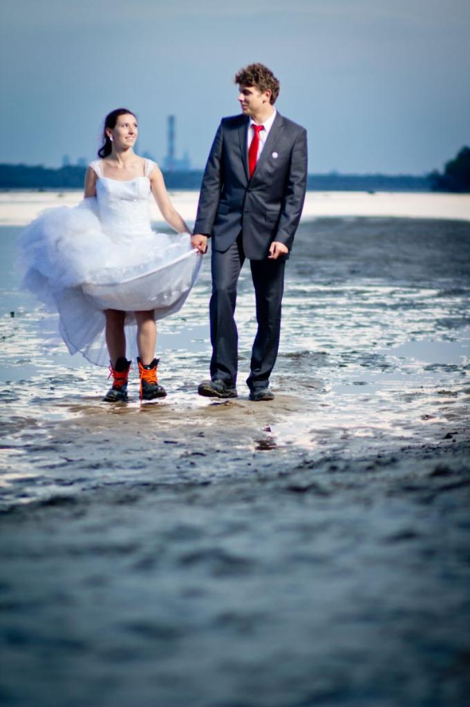 Para młoda na sesji plenerowej: panna młoda w białej sukni i pomarańczowych butach trekkingowych, pan młody w garniturze z czerwoną muchą, w tle krajobraz z kominami.