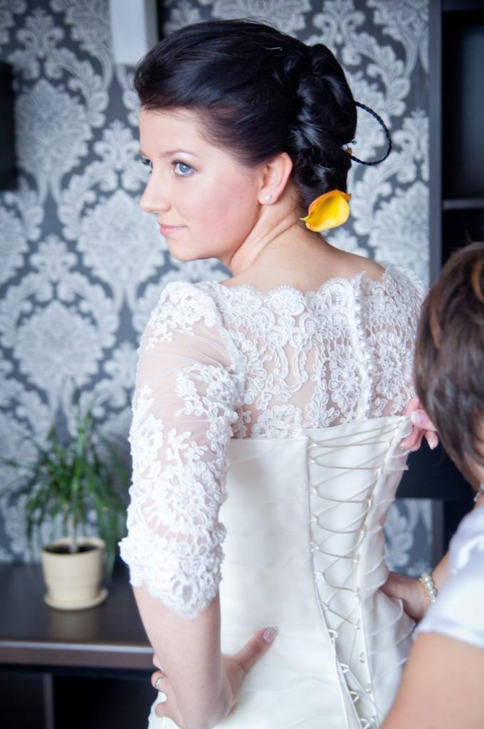 Panna młoda w białej sukni z koronkowymi rękawami, z żółtym kwiatem wpiętym we włosy, widok z tyłu podczas dopinania sukni przez drugą osobę, wzorzysta tapeta w tle.