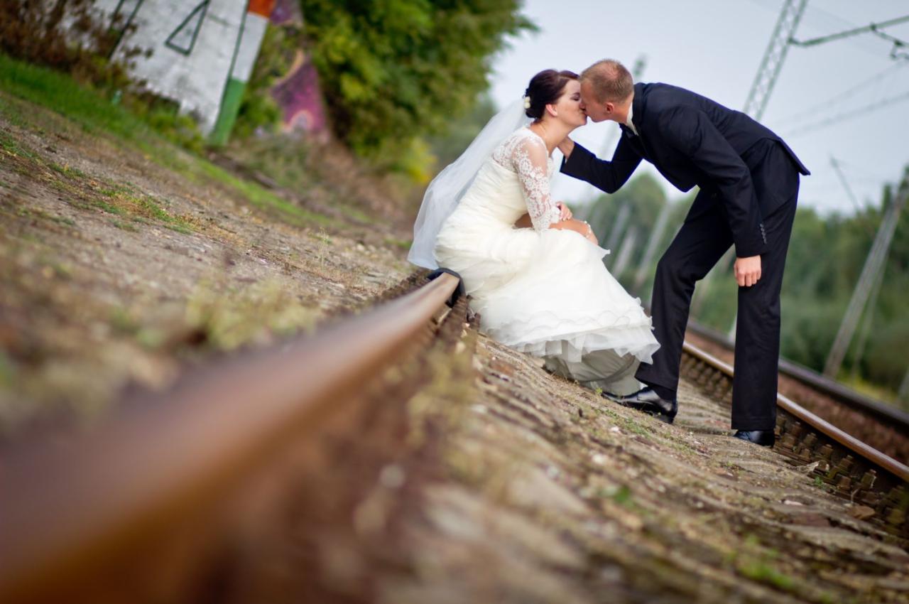 Pocałunek nowożeńców na torach kolejowych; panna młoda w białej sukni z koronkowymi rękawami siedzi na szynie, pan młody w czarnym garniturze pochyla się, tło z zielenią i słupami elektrycznymi.