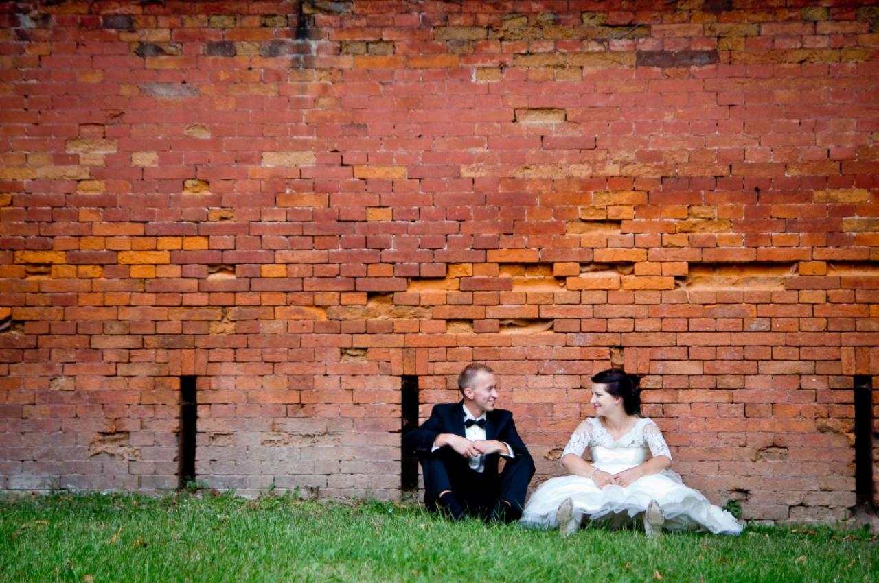 Para młoda siedzi na trawie pod ceglaną ścianą, ona w białej sukni z koronkowymi rękawami, on w czarnym smokingu z muszką, oboje patrzą na siebie z uśmiechem.