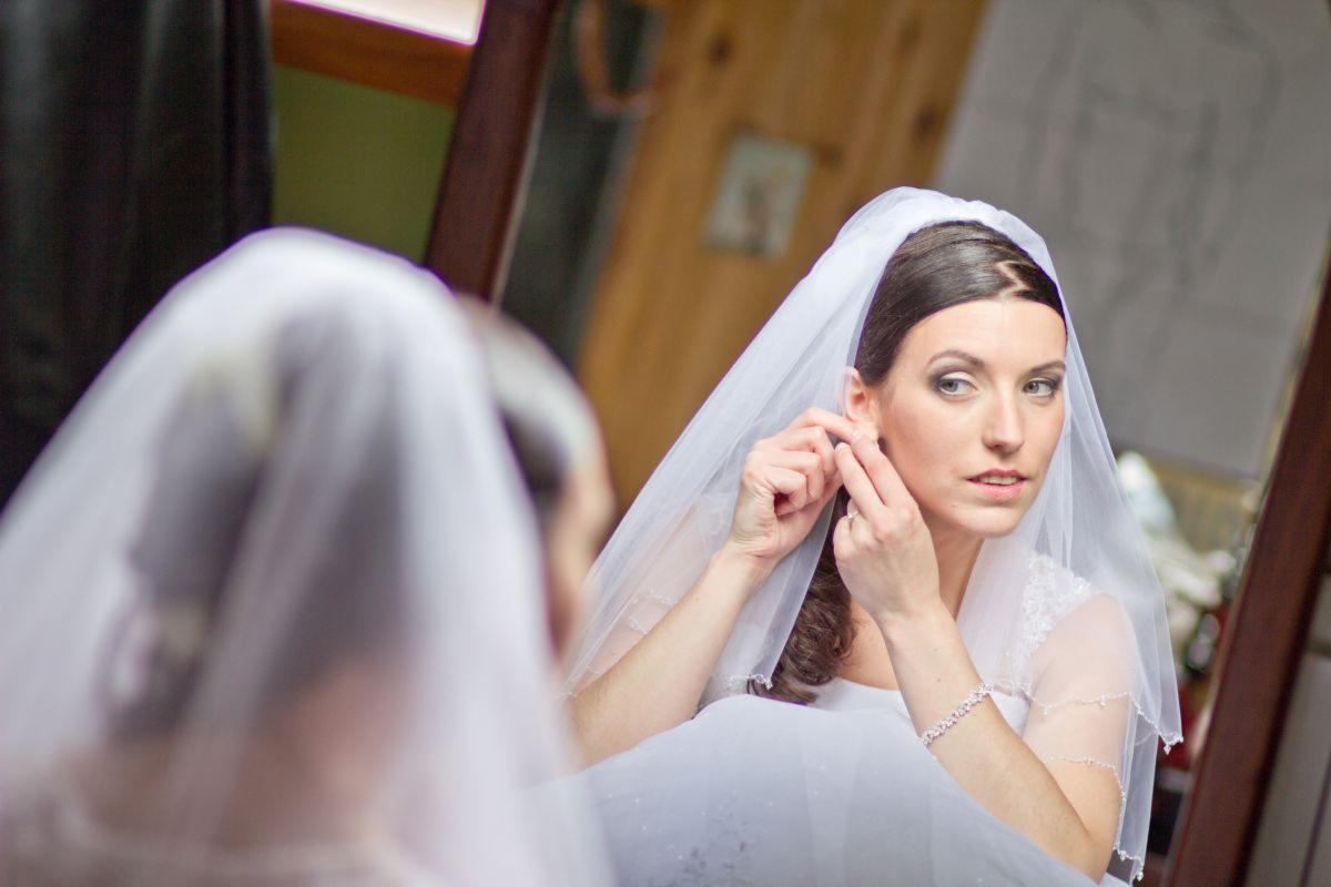 Panna młoda w welonie, patrząc w lustro, zakłada kolczyk przed ślubem.