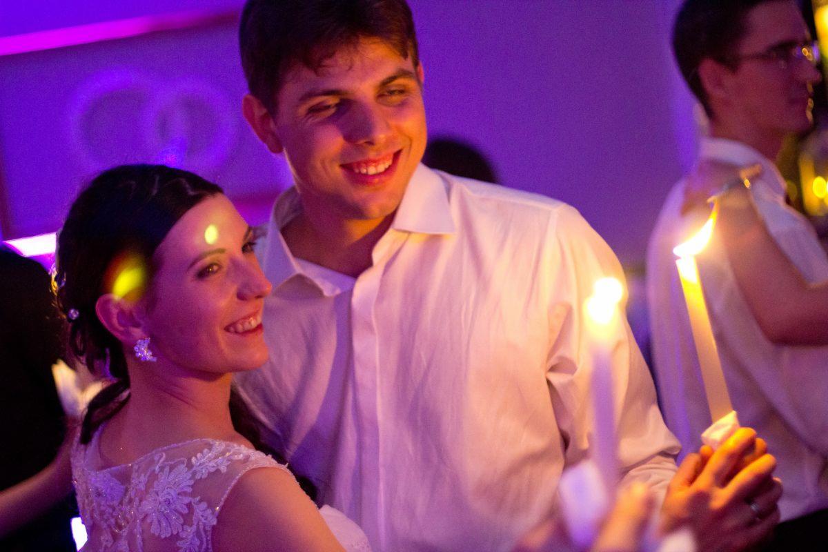 Uśmiechnięta para młoda podczas uroczystości ślubnej, w tle osoby trzymające zapalone świece, fioletowe oświetlenie.