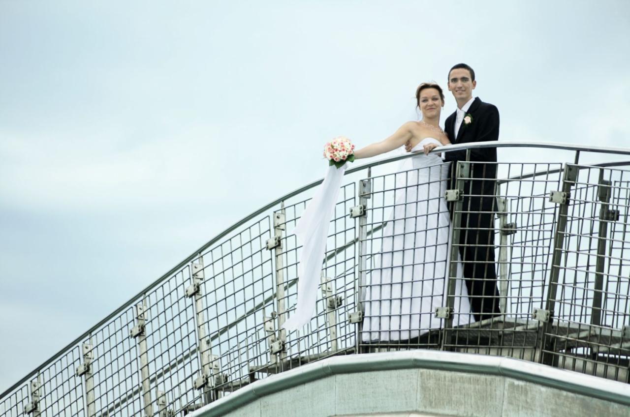 Para młoda w strojach ślubnych pozuje na metalowym moście z widokiem na pochmurne niebo. Panna młoda trzyma bukiet z długą, zwiewną wstążką.
