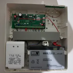 Wnętrze skrzynki alarmowej z centralą Satel Perfecta, transformatorem Pulsar i akumulatorem MW Power, widoczne diody LED i okablowanie.