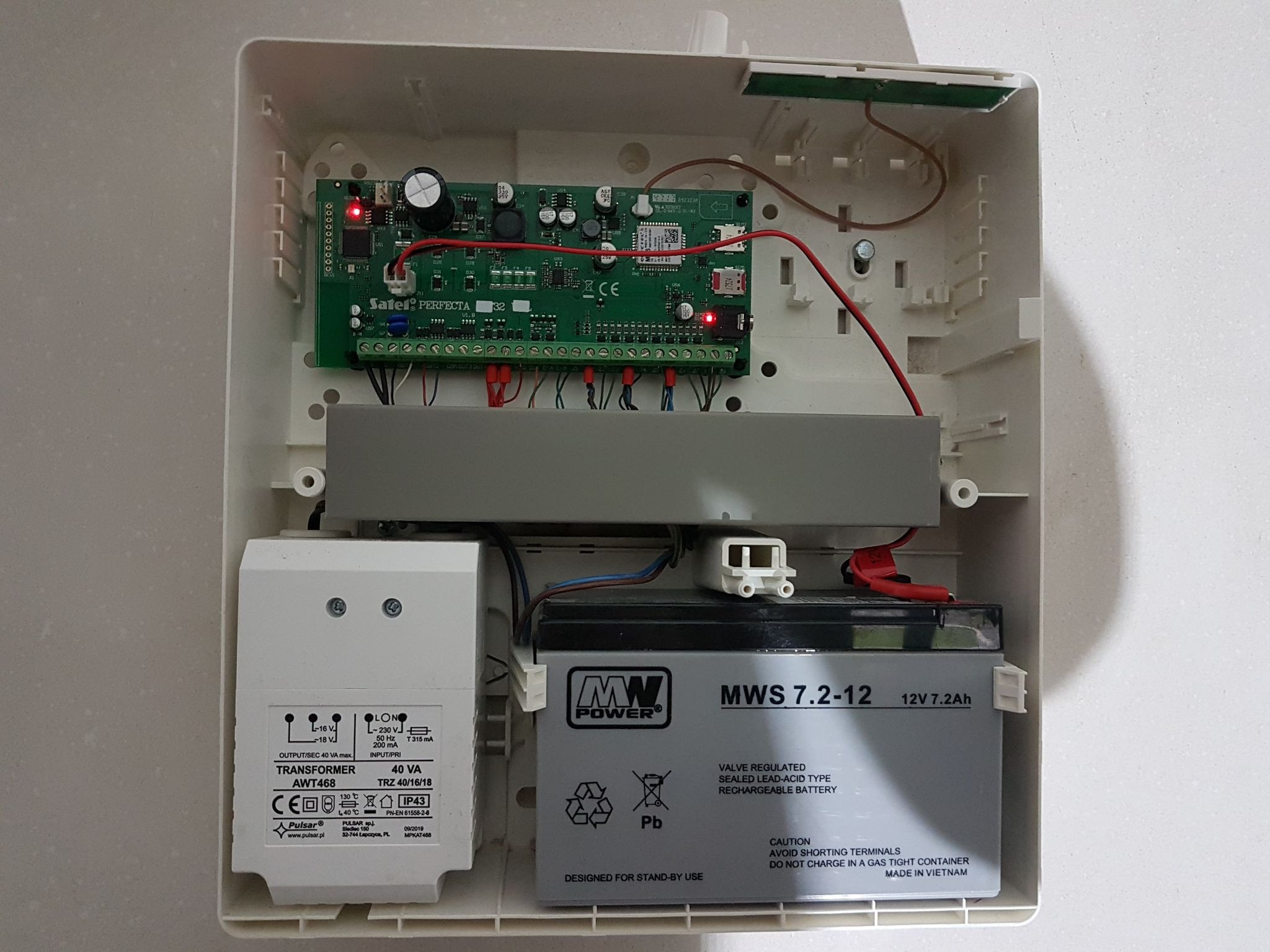 Wnętrze skrzynki alarmowej z centralą Satel Perfecta, transformatorem Pulsar i akumulatorem MW Power, widoczne diody LED i okablowanie.