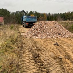 Wo-Kaz - Ciężarówka z wywrotką stoi za stertą potłuczonych cegieł na placu budowy, widoczne ślady opon na błotnistym podłożu i budynki w tle.