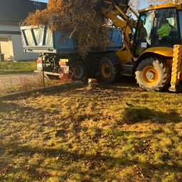 Wo-Kaz - Żółta koparko-ładowarka załadowuje gałęzie z liśćmi do niebieskiej ciężarówki na tle budynku mieszkalnego i błękitnego nieba.