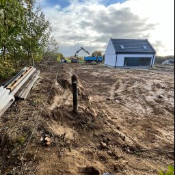 Wo-Kaz - Przygotowanie terenu pod budowę domu jednorodzinnego: wyrównana działka z widocznym wykopem, rurą wystającą z ziemi, stertą desek, sznurkiem wyznaczającym granice, koparką i wywrotką w tle,...