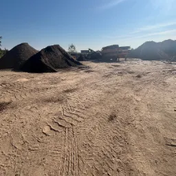 Kielno: Stosy ciemnej ziemi ogrodowej i maszyna do przesiewania na placu, ślady opon na piaszczystym podłożu, błękitne niebo.