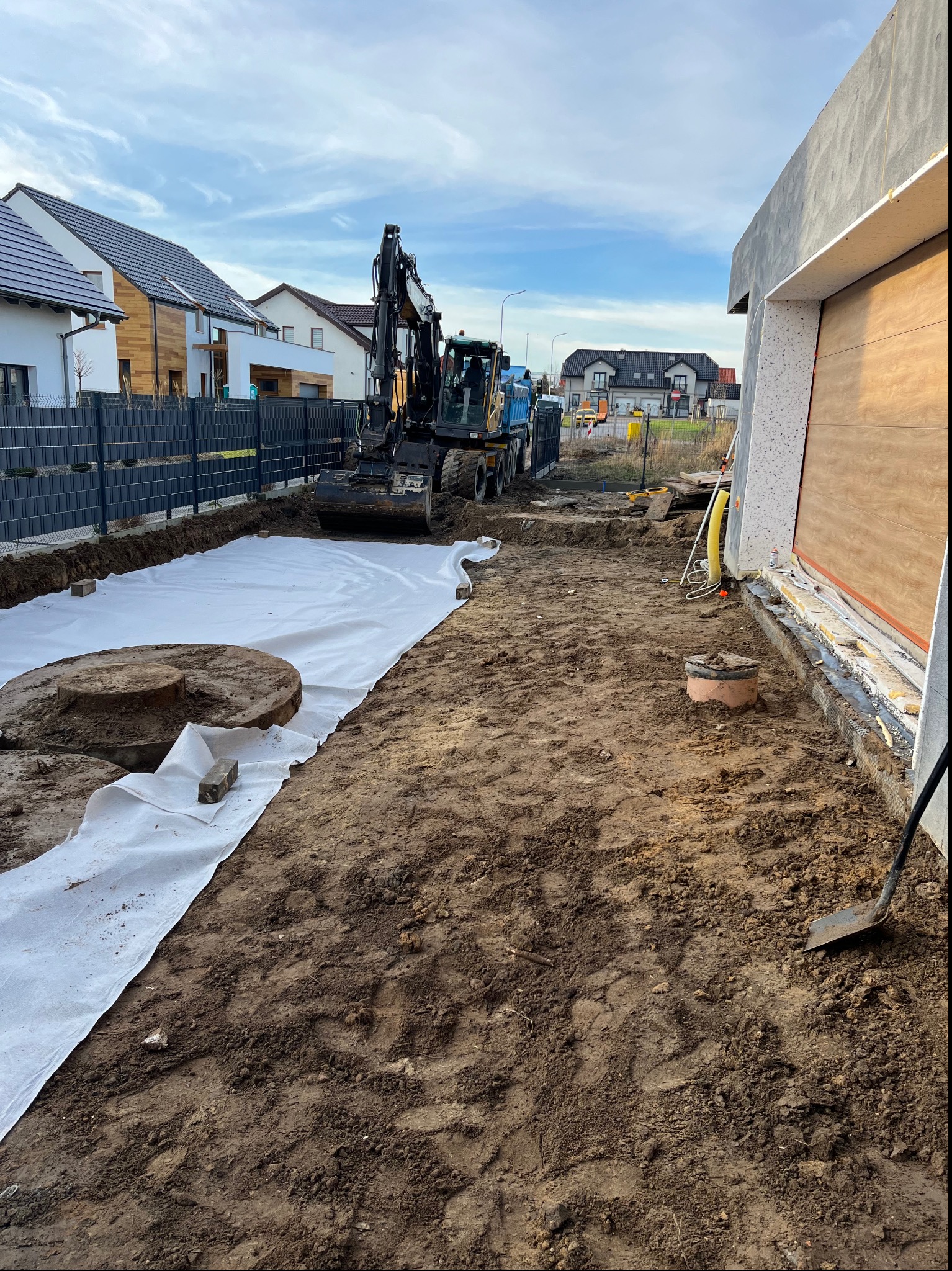 Przygotowanie terenu pod budowę: wykopany rów wyłożony białą geowłókniną, w tle koparka i wywrotka, widoczne elementy kanalizacji i fundamentów budynku.