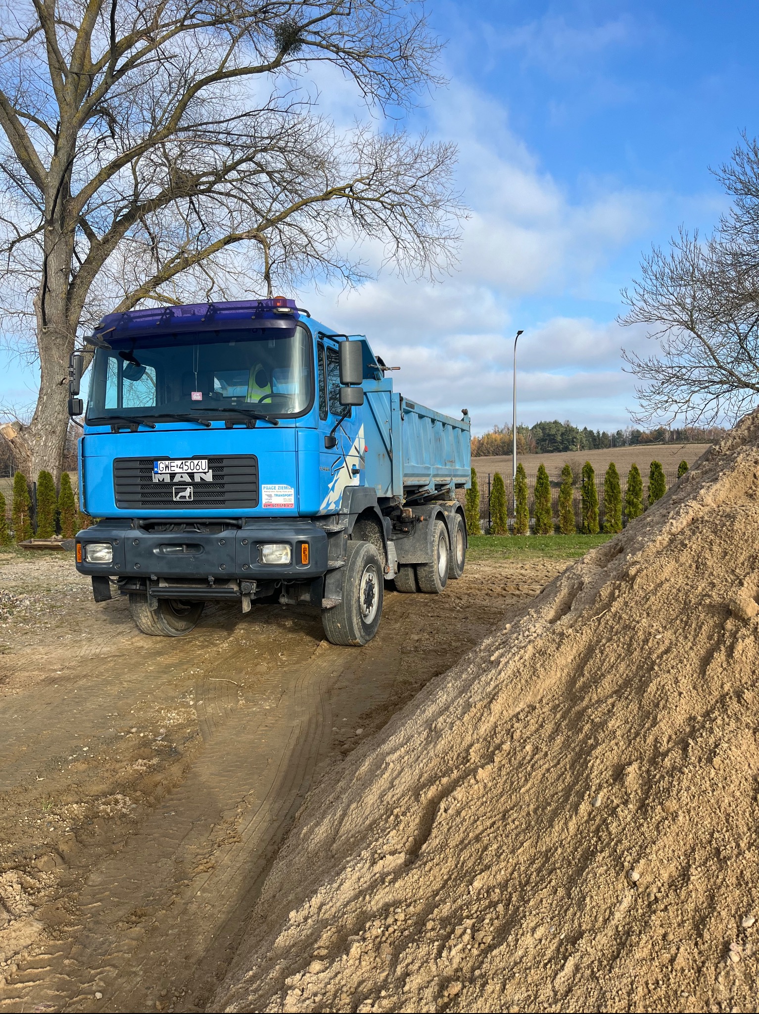 Niebieska ciężarówka MAN z wywrotką stoi na ziemi obok pryzmy piasku, w tle drzewa bez liści i rząd tuj na granicy pola.