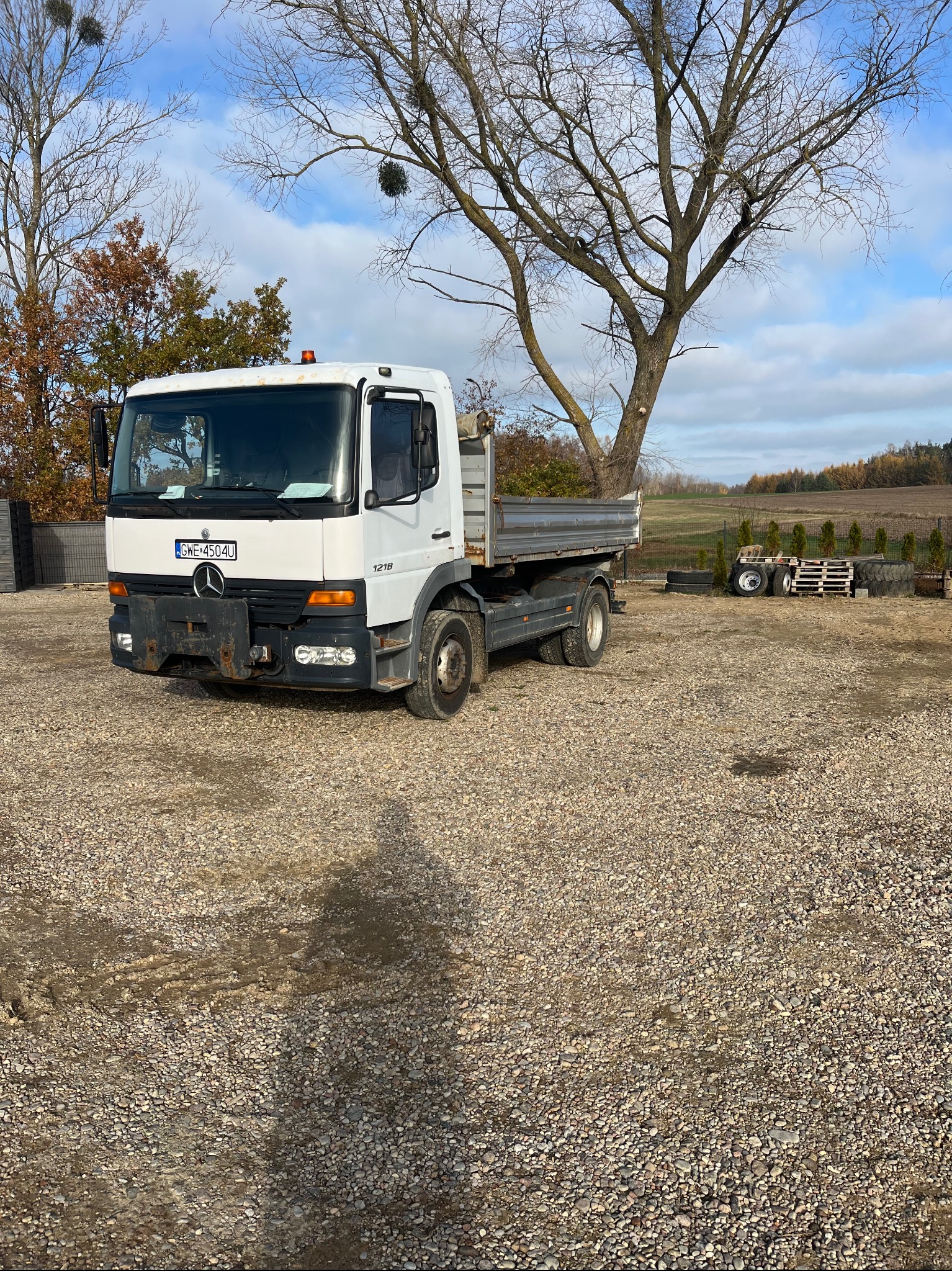 Biała ciężarówka Mercedes-Benz 1218 z otwartą skrzynią ładunkową, zaparkowana na żwirowym placu z widokiem na pole i drzewa w tle.