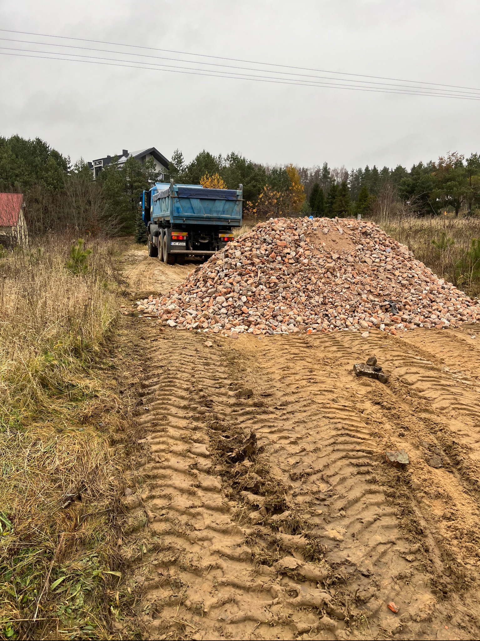 Ciężarówka z wywrotką stoi za stertą potłuczonych cegieł na placu budowy, widoczne ślady opon na błotnistym podłożu i budynki w tle.