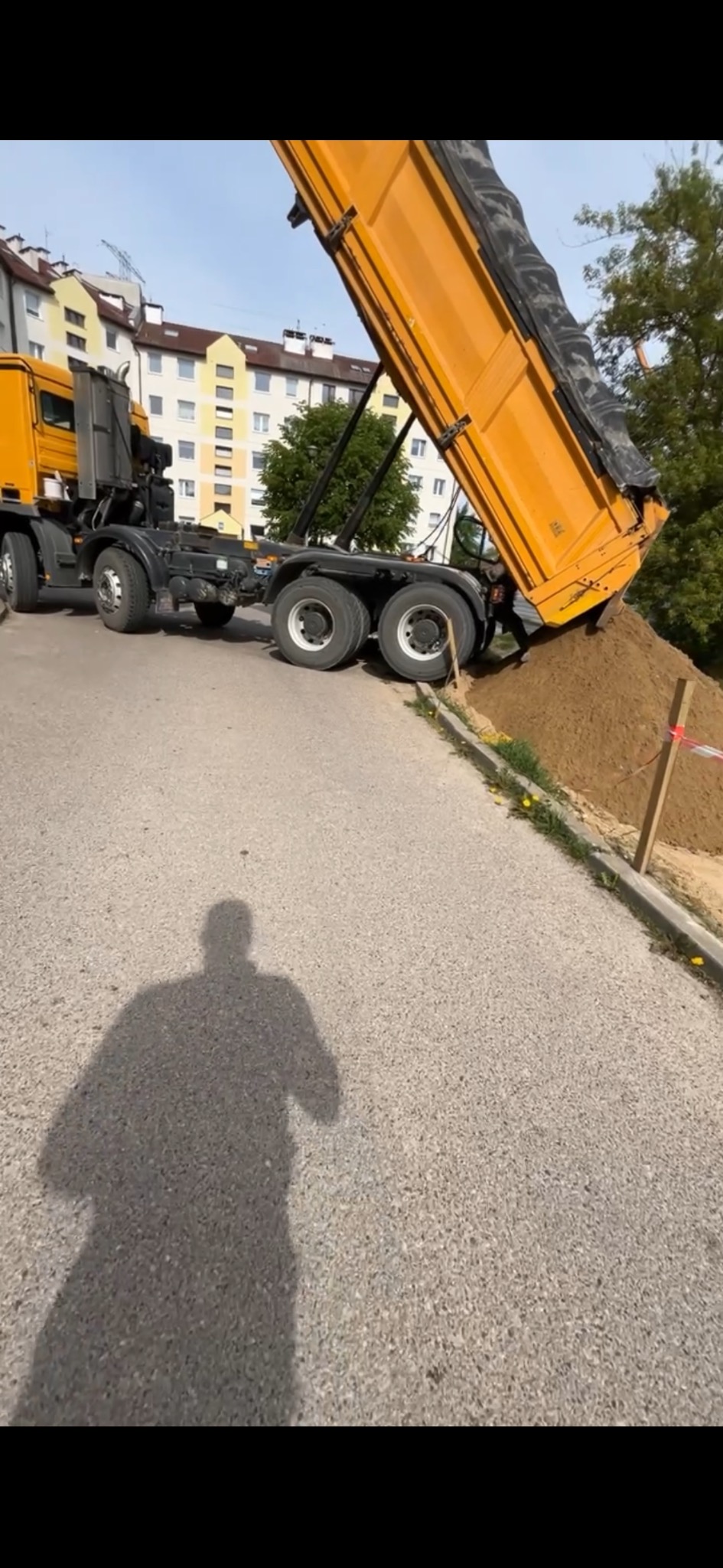 Żółta wywrotka wysypuje ziemię na pobocze drogi w słoneczny dzień, widoczny cień fotografa.