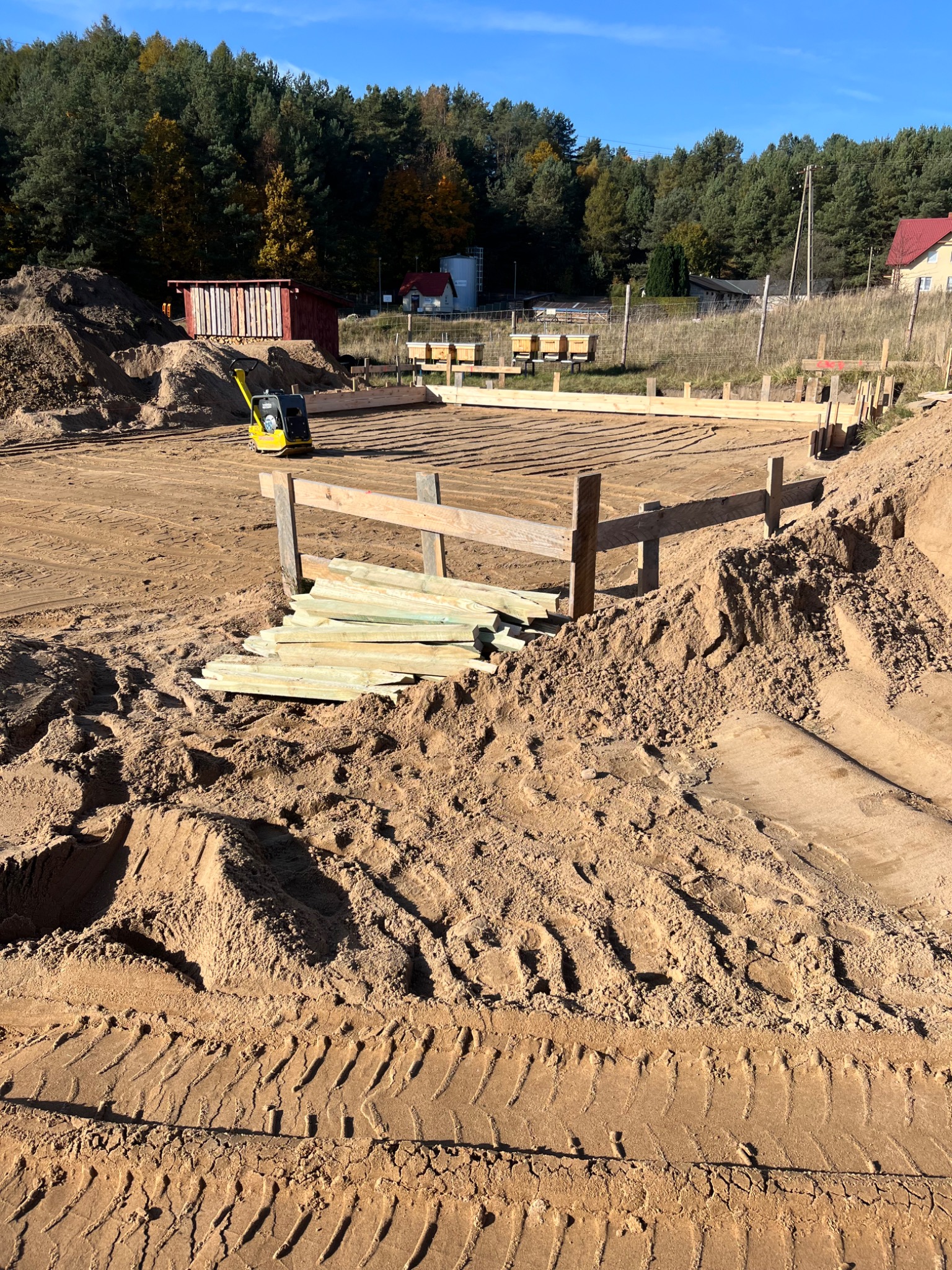 Przygotowany teren pod fundamenty z widocznymi drewnianymi szalunkami, stertą desek i małą zagęszczarką gruntu w tle, na piaszczystym terenie z widocznymi śladami opon.