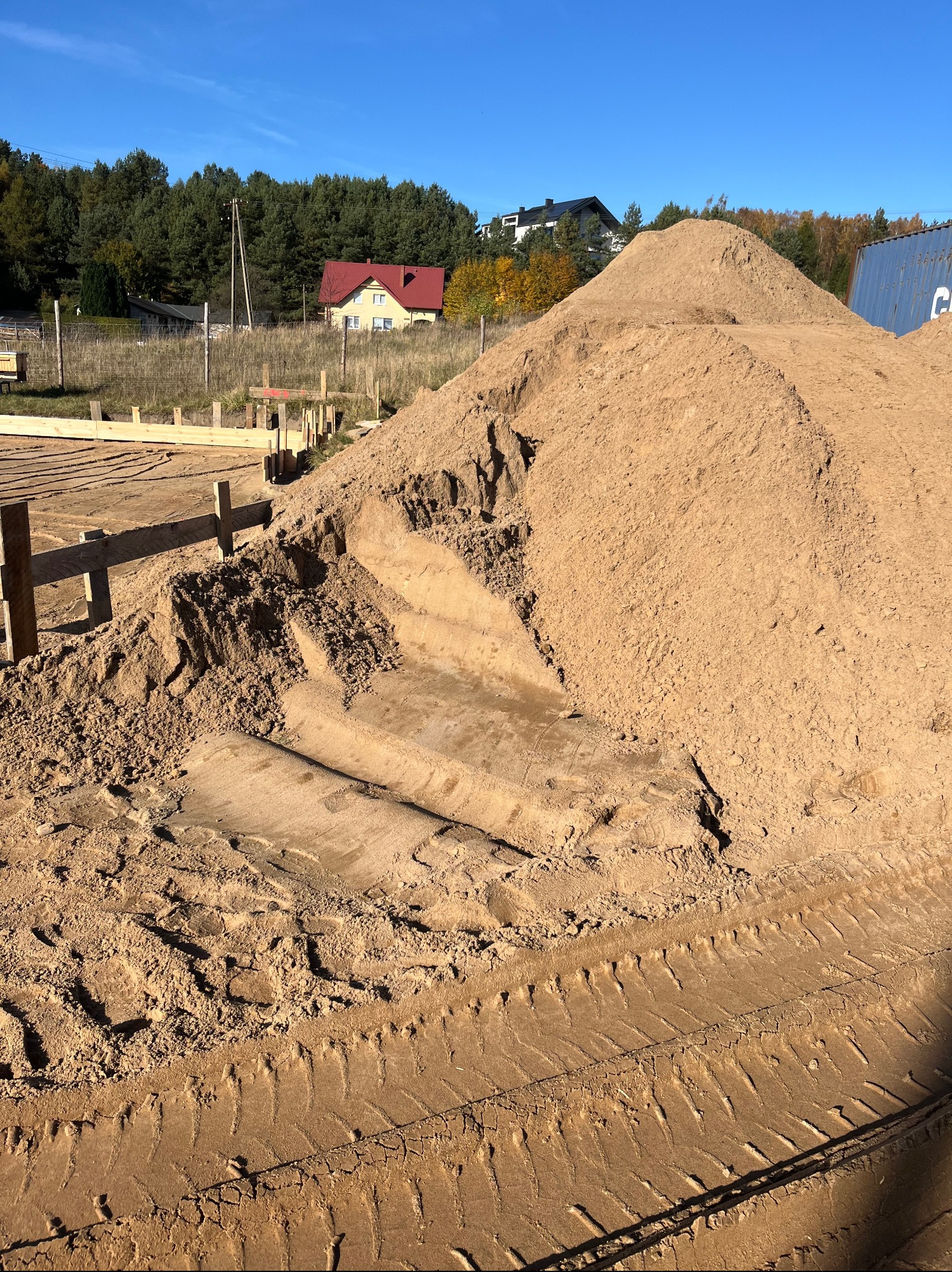 Kupa piasku z widocznymi śladami opon na placu budowy z drewnianą konstrukcją fundamentów w tle i zabudowaniami mieszkalnymi