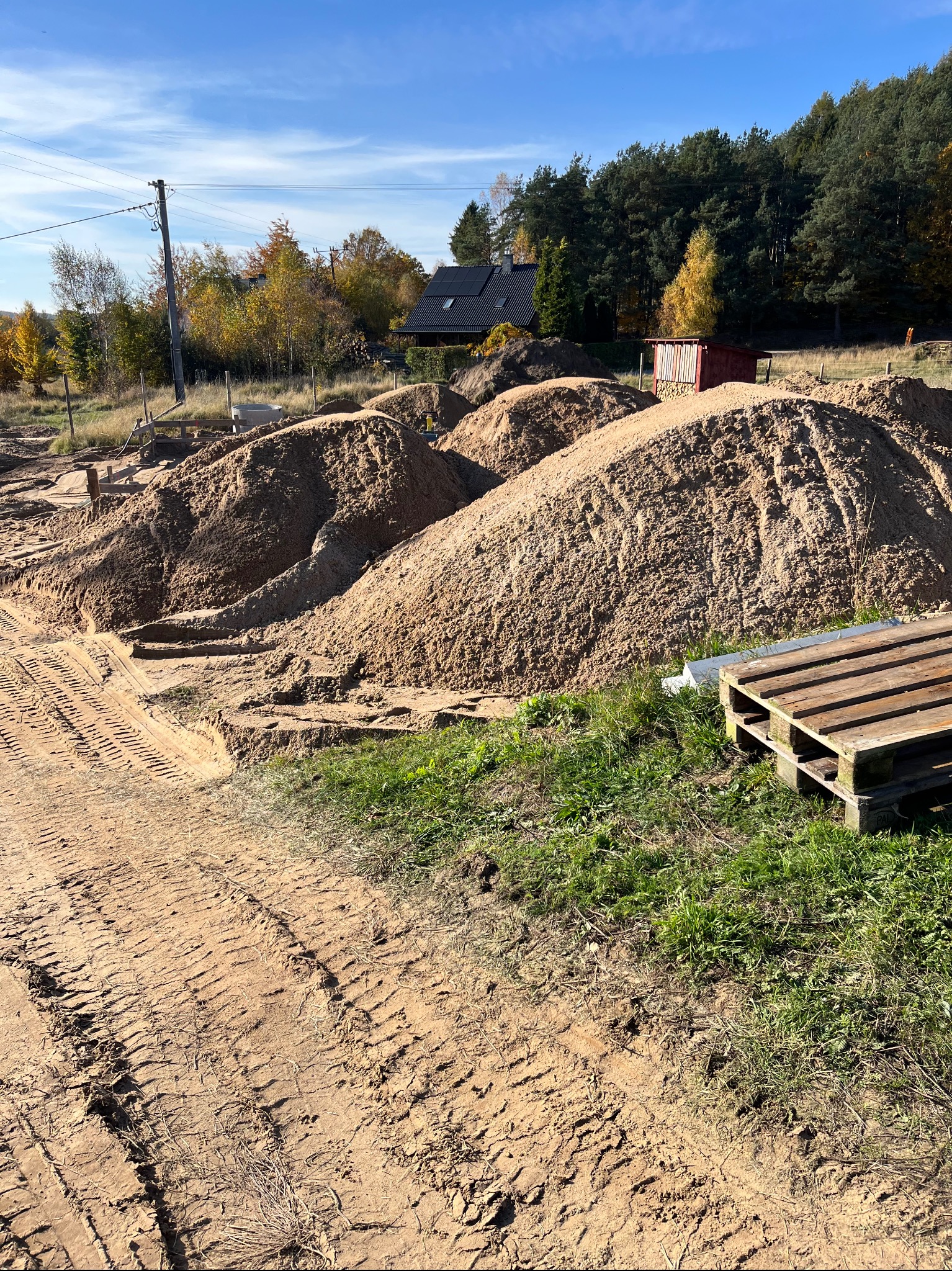 Stosy piasku na placu budowy z widocznymi śladami opon i drewnianymi paletami, w tle dom jednorodzinny i drzewa.