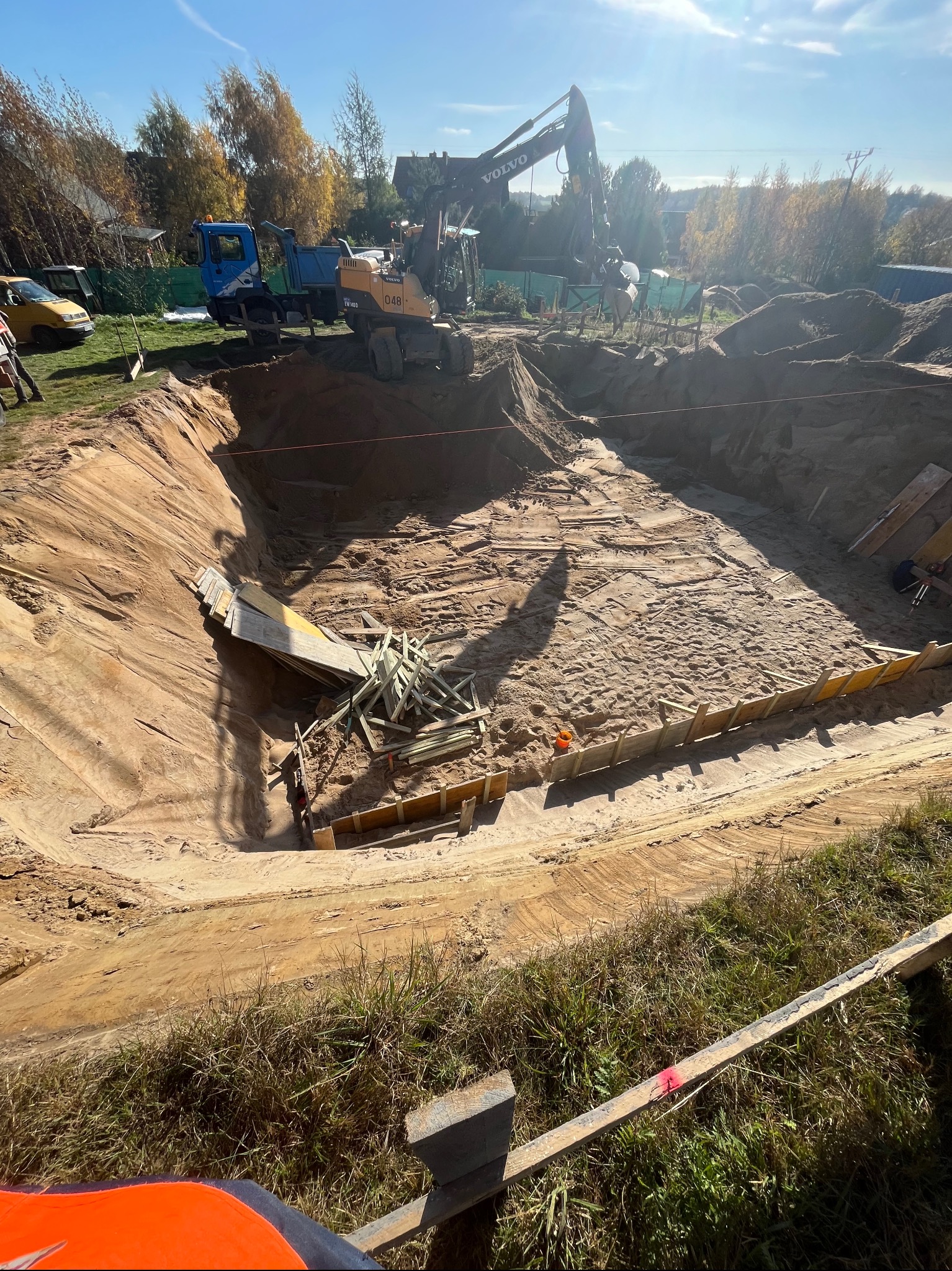 Głęboki wykop pod budowę z widocznymi deskami szalunkowymi, stertą drewna wewnątrz i pracującą koparką Volvo w tle, na zewnątrz wykopu zaparkowany samochód dostawczy i ciężarówka.
