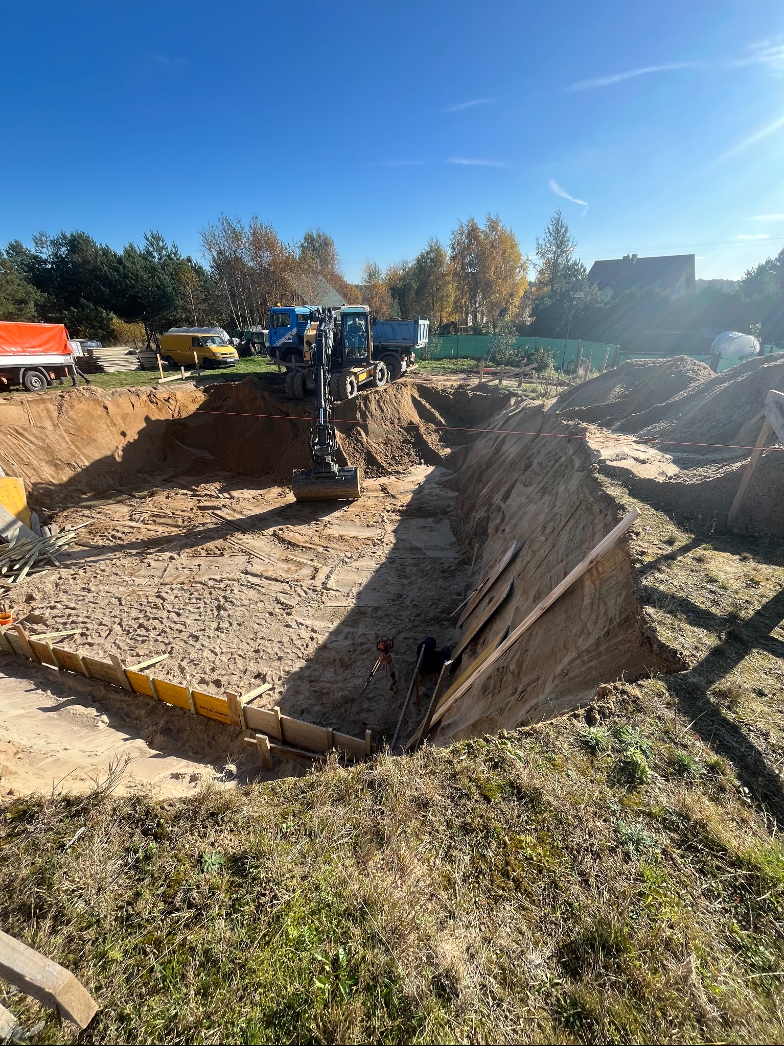 Głęboki wykop pod fundamenty z deskowaniem i pracującą koparką, na tle błękitnego nieba oraz zieleni drzew i zabudowań.