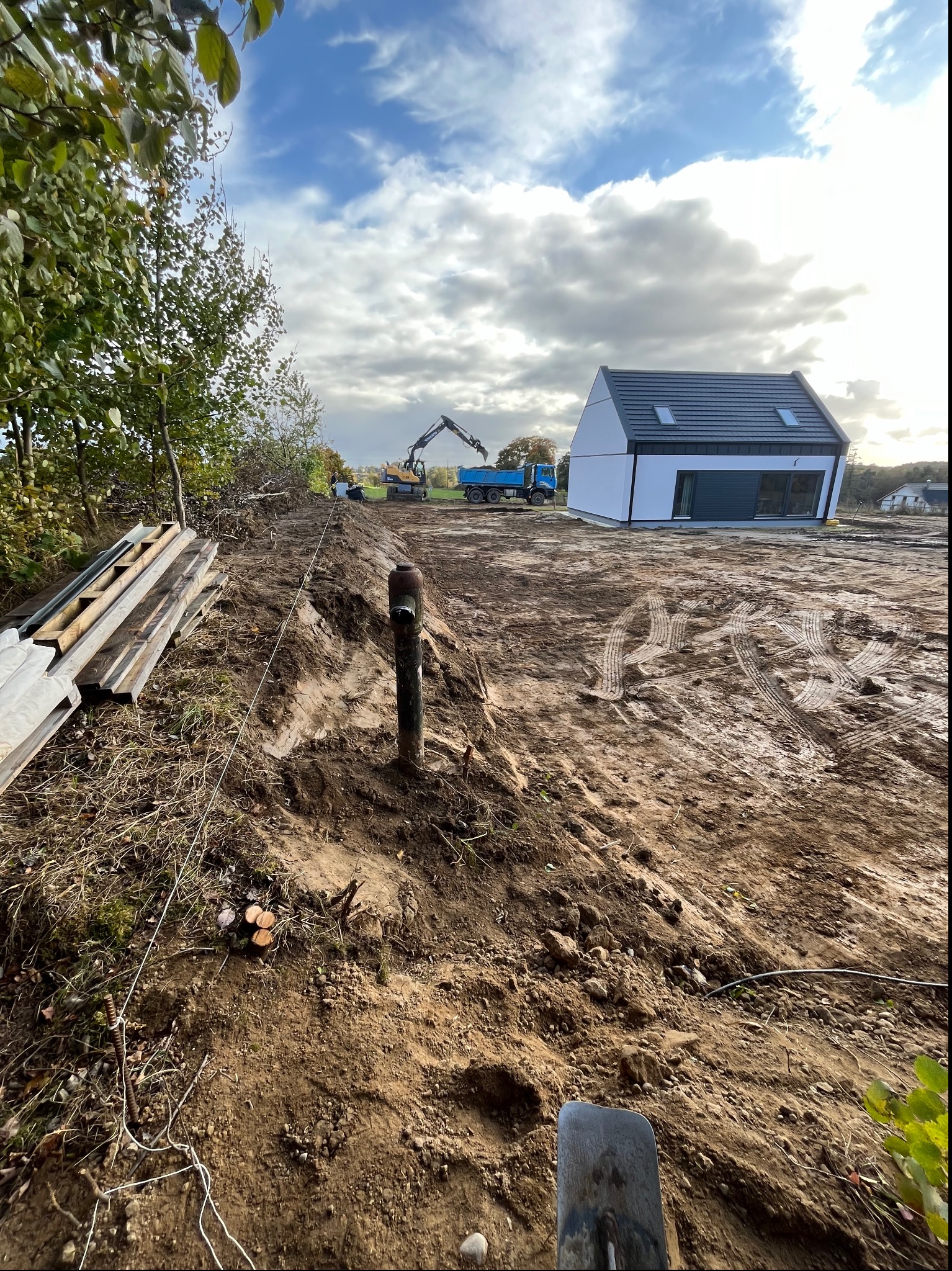 Przygotowanie terenu pod budowę domu jednorodzinnego: wyrównana działka z widocznym wykopem, rurą wystającą z ziemi, stertą desek, sznurkiem wyznaczającym granice, koparką i wywrotką w tle,...